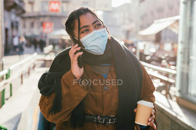 Italia, la mujer en la máscara de la cara que sostiene la taza desechable y el uso de teléfono inteligente - foto de stock