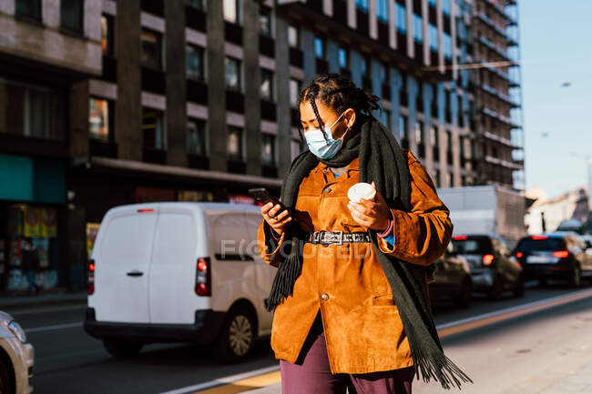 Італія, Жінка в масці, яка тримає смартфон і одноразову чашку на вулицях міста — стокове фото