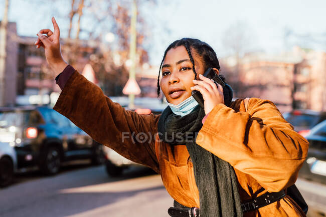 Italy, Woman with face mask hailing cab and talking on mobile phone on street — Stock Photo