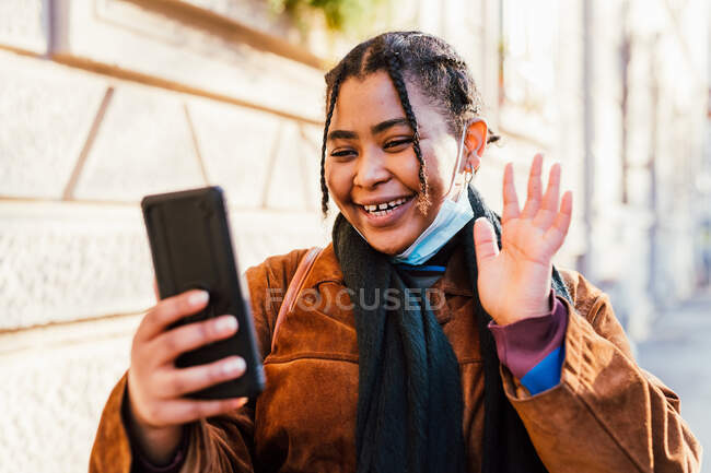 Italy, Young woman with face mask smiling and waving to smart phone — Stock Photo