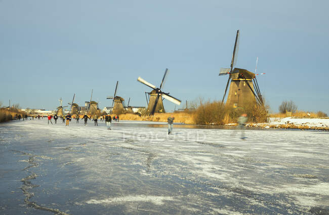 Nederland, Zuid-Holland, Kinderdijk, Gente patinando sobre hielo cerca de molinos de viento - foto de stock