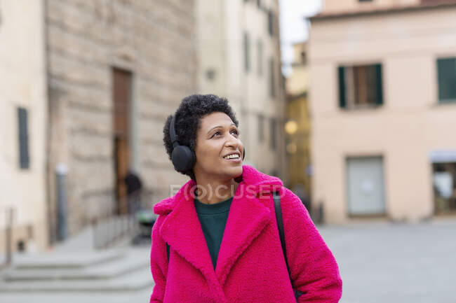Italia, Toscana, Pistoia, Mujer de abrigo rosa y auriculares caminando por la ciudad - foto de stock