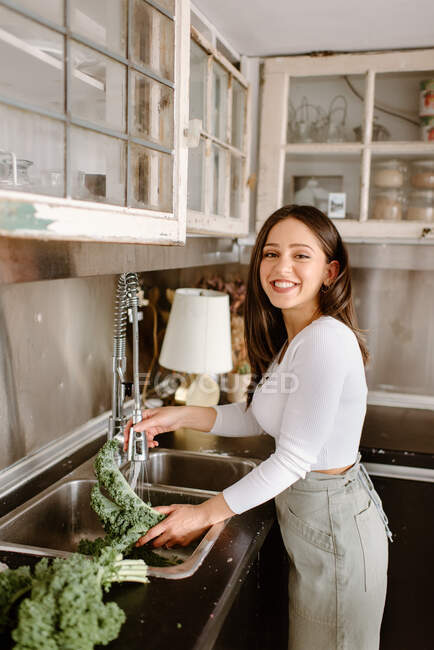 Souriant jeune femme laver le chou frisé dans l'évier de cuisine — Photo de stock