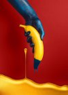 Hand in blue paint holding banana on red background — Stock Photo