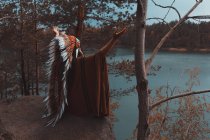 Portrait arrière de femme en casque amérindien assis sur la pierre près de la rivière avec les mains tendues — Photo de stock