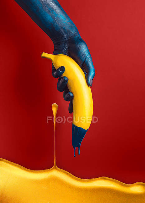 Hand in blue paint holding banana on red background — Stock Photo
