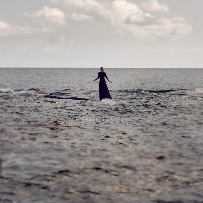 Retrato trasero de mujer en vestido negro de pie en la superficie del agua en el mar - foto de stock