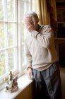 Старший чоловік з долонею на обличчі стоїть біля вікна в домашньому інтер'єрі . — стокове фото