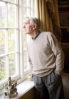 Lonely senior man looking through window in home interior. — Stock Photo