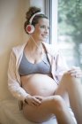 Беременная женщина в нижнем белье слушает музыку в наушниках на подоконнике . — стоковое фото