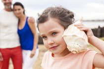 Young girl listening to seashell with parents in background. — Stock Photo