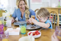 Junge gießt Ahornsirup auf Pfannkuchen am Esstisch mit Mutter. — Stockfoto