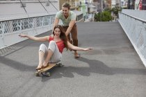 Donna seduta sullo skateboard con uomo che spinge sulla strada . — Foto stock
