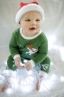 Baby boy in Christmas pajamas playing with fairy lights. — Stock Photo