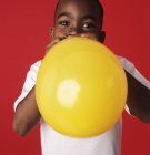 Boy blowing up yellow balloon on red background. — Stock Photo