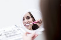Paciente feminina escovando os dentes enquanto olha no espelho . — Fotografia de Stock