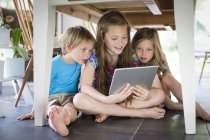 Three siblings under table using digital tablet. — Stock Photo
