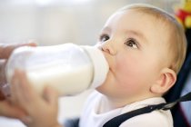 Female hand helping baby boy drinking bottle of milk in safety chair. — Stock Photo