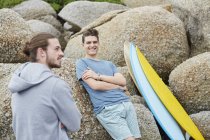 Young men leaning on rocks with surfboard. — Stock Photo