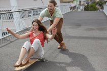 Woman sitting on skateboard with man pushing on street. — Stock Photo