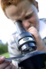 Garçon rousse utilisant un microscope léger pour étudier sur prairie . — Photo de stock