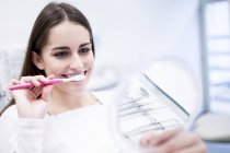 Paciente feminina escovando os dentes enquanto olha no espelho . — Fotografia de Stock