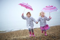Two preschooler girls running on beach with pink umbrellas and holding hands. — Stock Photo