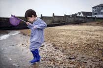 Menino pré-escolar em botas de borracha na praia segurando rede de pesca . — Fotografia de Stock