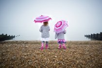 Zwei Vorschulmädchen, die am Strand spazieren gehen und rosa Regenschirme in der Hand halten. — Stockfoto