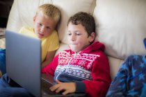 Dois meninos usando laptop enquanto sentados no sofá em casa . — Fotografia de Stock