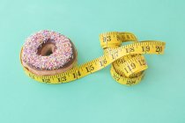 Doughnut with sprinkles and yellow tape measure against green background. — Stock Photo