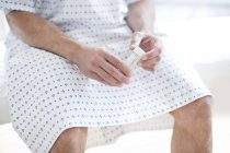 Male patient in hospital gown sitting on bed holding urine sample container. — Stock Photo