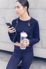 Женщина в спортивной одежде держит бутылку воды и смартфон на улице . — стоковое фото