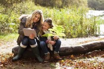Mother using digital tablet with daughter on log in forest. — Stock Photo