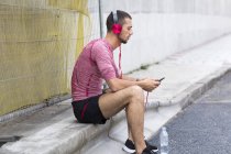 Hombre sentado en la calle bordillo y escuchar música en el teléfono inteligente . - foto de stock