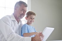 Дедушка и внук используют цифровой планшет в помещении и улыбаются — стоковое фото
