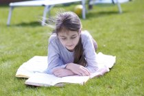 Preteen girl lying on blanket on green meadow in garden and reading book. — Stock Photo