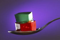 Cubes of red and green jelly on spoon on purple background. — Stock Photo