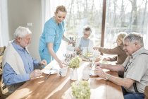 Female care worker serving senior adults at dinner table in care home. — Stock Photo