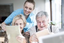Cuidador alegre ajudando casal sênior usando tablets digitais em casa de cuidados . — Fotografia de Stock
