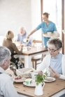 Senior adults eating breakfast in care home while caregiver serving. — Stock Photo