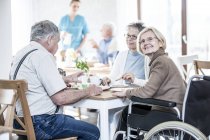 Adultes âgés dînant dans une maison de soins tandis que la femme en fauteuil roulant regarde à la caméra . — Photo de stock