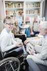 Femme âgée avec tasse en fauteuil roulant assis avec des amis masculins et infirmière dans la maison de soins . — Photo de stock