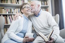Senior man kissing woman on cheek and embracing while reading book and holding tea. — Stock Photo