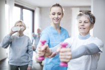 Female physiotherapist helping senior woman holding hand weights with woman drinking in background. — Stock Photo