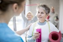 Старша жінка з пляшкою розмовляє з фізіотерапевтом у класі фізичних вправ . — стокове фото
