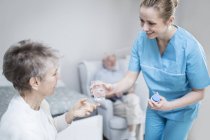 Care worker giving senior woman medication in care home with man in background. — Stock Photo