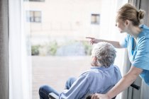 Homme âgé en fauteuil roulant avec travailleur de soins regardant par la fenêtre . — Photo de stock