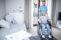 Enfermera empujando a una mujer mayor en silla de ruedas en un hogar de cuidados . - foto de stock