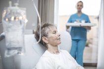 Mujer mayor con cánula nasal y bolsa IV y bandeja de sujeción de enfermera en segundo plano, primer plano . - foto de stock